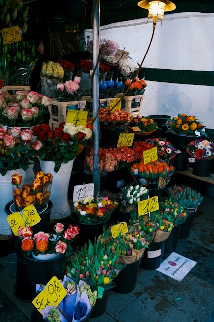 A vibrant display of fresh flower bouquets arranged in black plastic buckets and white vases at a flower stall, featuring a variety of colorful blooms including pink roses, red roses, yellow and orang