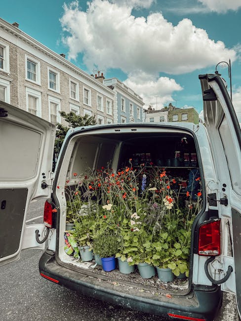 The open back of a white delivery van filled with potted flowers and plants, including bright red poppies, white cosmos, and various green foliage, parked on a street with classic Victorian-style buil