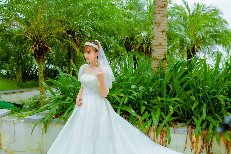 A young woman dressed in a white wedding gown with puffed sleeves and a fitted bodice, holding a bouquet of fresh flowers, stands outdoors amidst lush green foliage, including palm trees and broad-lea